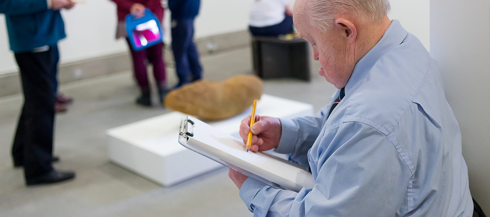 Blue Room artist in the foreground is drawing on a sketch pad while other members browse the gallery in the background.