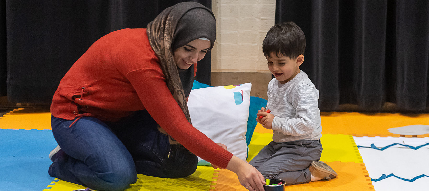 A mother and child play on a blue, yellow and orange coloured floor.