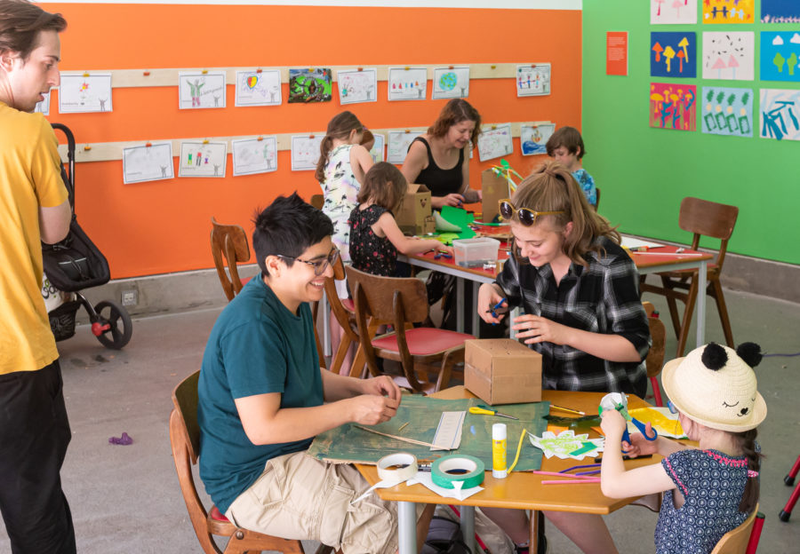 Two families, the one on the background features one adult and three children, the one on the foreground features one adult two children. Each group are smiling as they cut and stick with a variety of art supplies on the table. The room has one orange wall and one green.