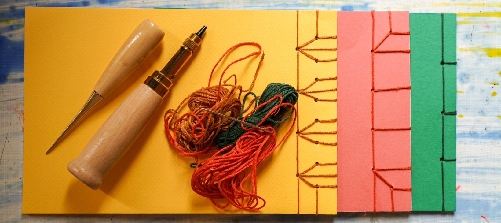 Image shows 3 notebooks laying on a table. The notebooks overlap on another, one green on the bottom, an orange overlapping this and finally a yellow one on the very top. Laying on the yellow notebook are book binding tools.