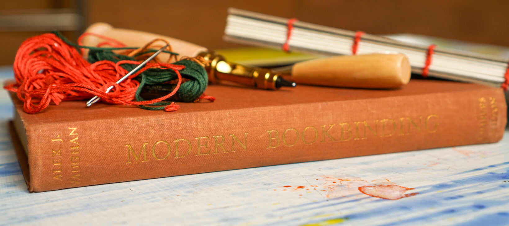Book binding tools lay on top of a book in a rust colour, that reads "Modern Bookbinding".