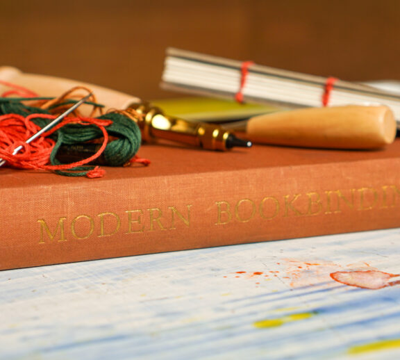 Book binding tools lay on top of a book in a rust colour, that reads "Modern Bookbinding".