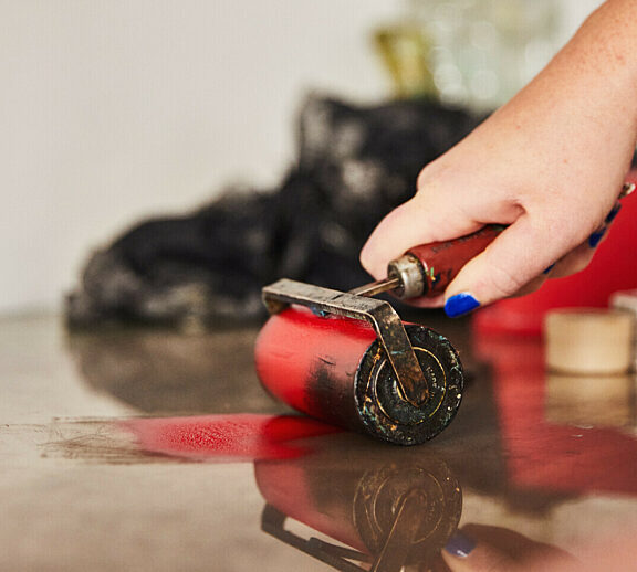 A hand is holding a roller that is spreading read paint onto a clear surface.