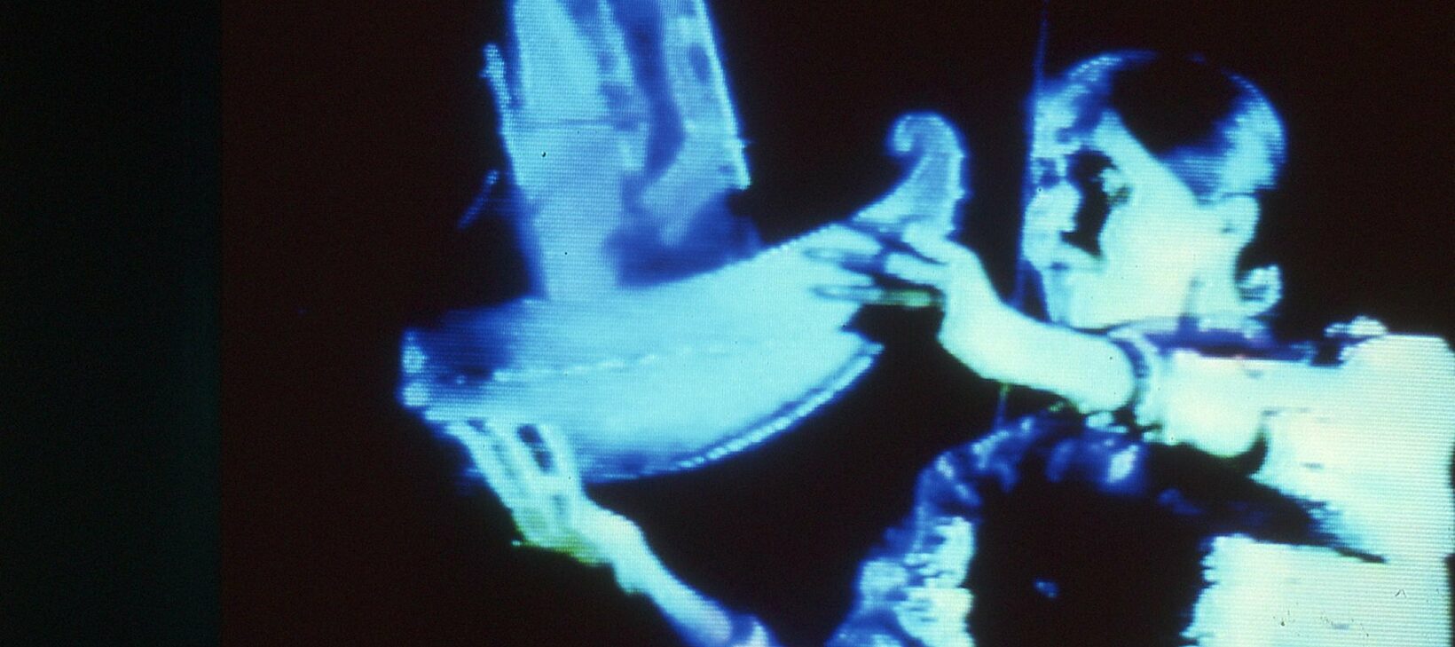 Images shoes a person holding a miniature sculpture of a boat, shrouded in blue light against a black background