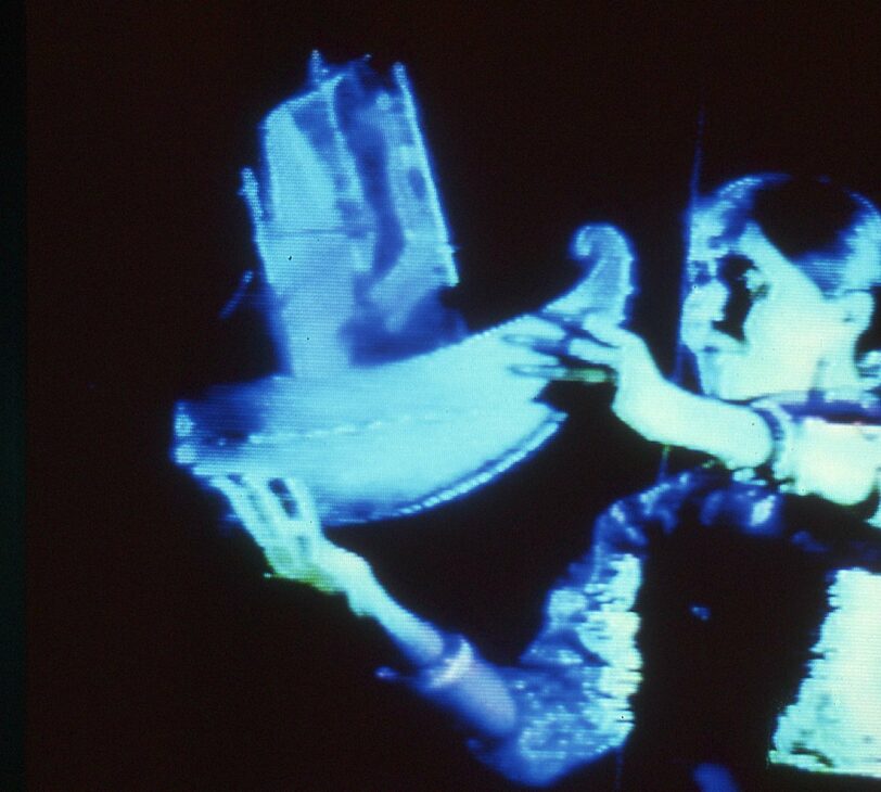 Images shoes a person holding a miniature sculpture of a boat, shrouded in blue light against a black background