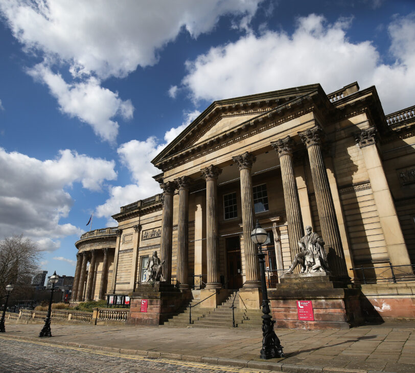 A photograph of a brown building with Greek-style columns in front of the entrance.