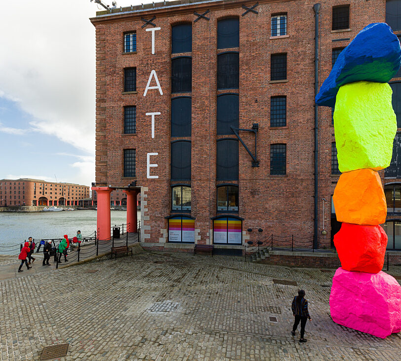 A photograph of a giant colourful sculpture, in front of a red brick building labelled 'Tate'.