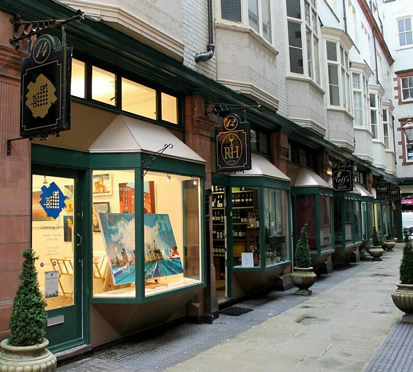 A photograph of a small gallery illuminated from the inside, with potted plants outside and a large canvas painting in the window.