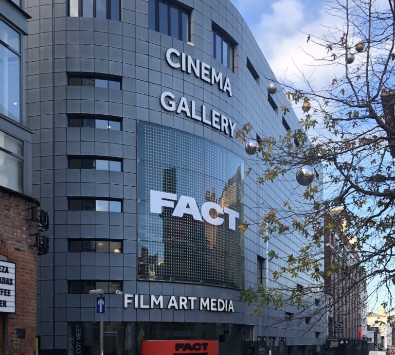 A photograph of a metallic building on the corner of a street, with a bright red doorframe and the words 'cinema, gallery, FACT, film art media' written in white.