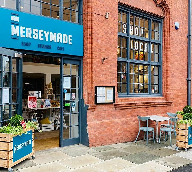 A photograph of a shop with a blue sign that says 'merseymade', a table and chairs outside, and some large planters with greenery in them.