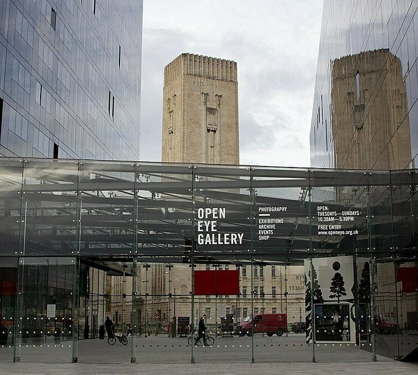A photograph of Open Eye Gallery, a glass building with the name written in white, and a tall brown building in the background.
