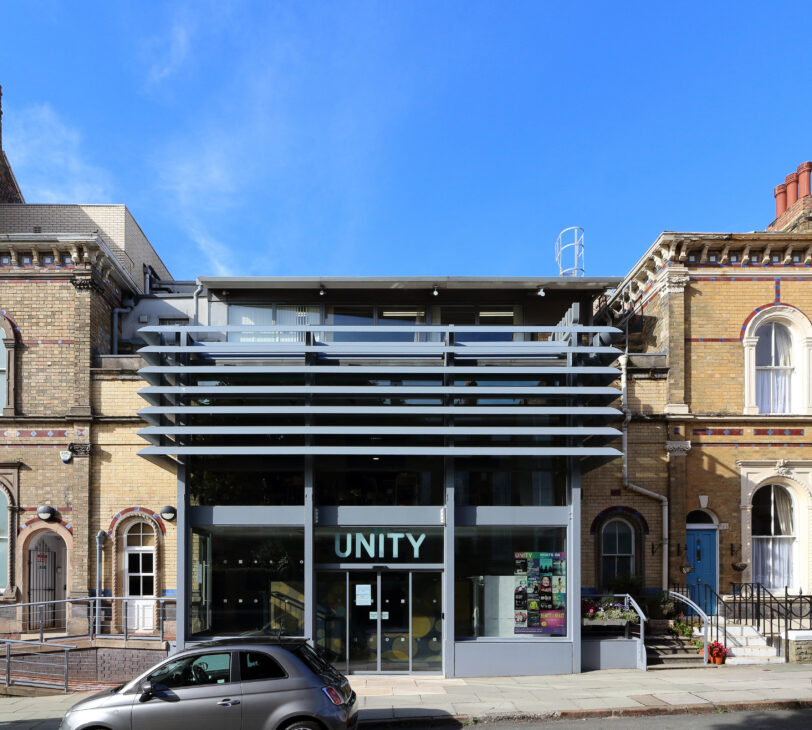 A photograph of a metal building in between two brown brick buildings, with the word 'Unity' written in white above the glass door.